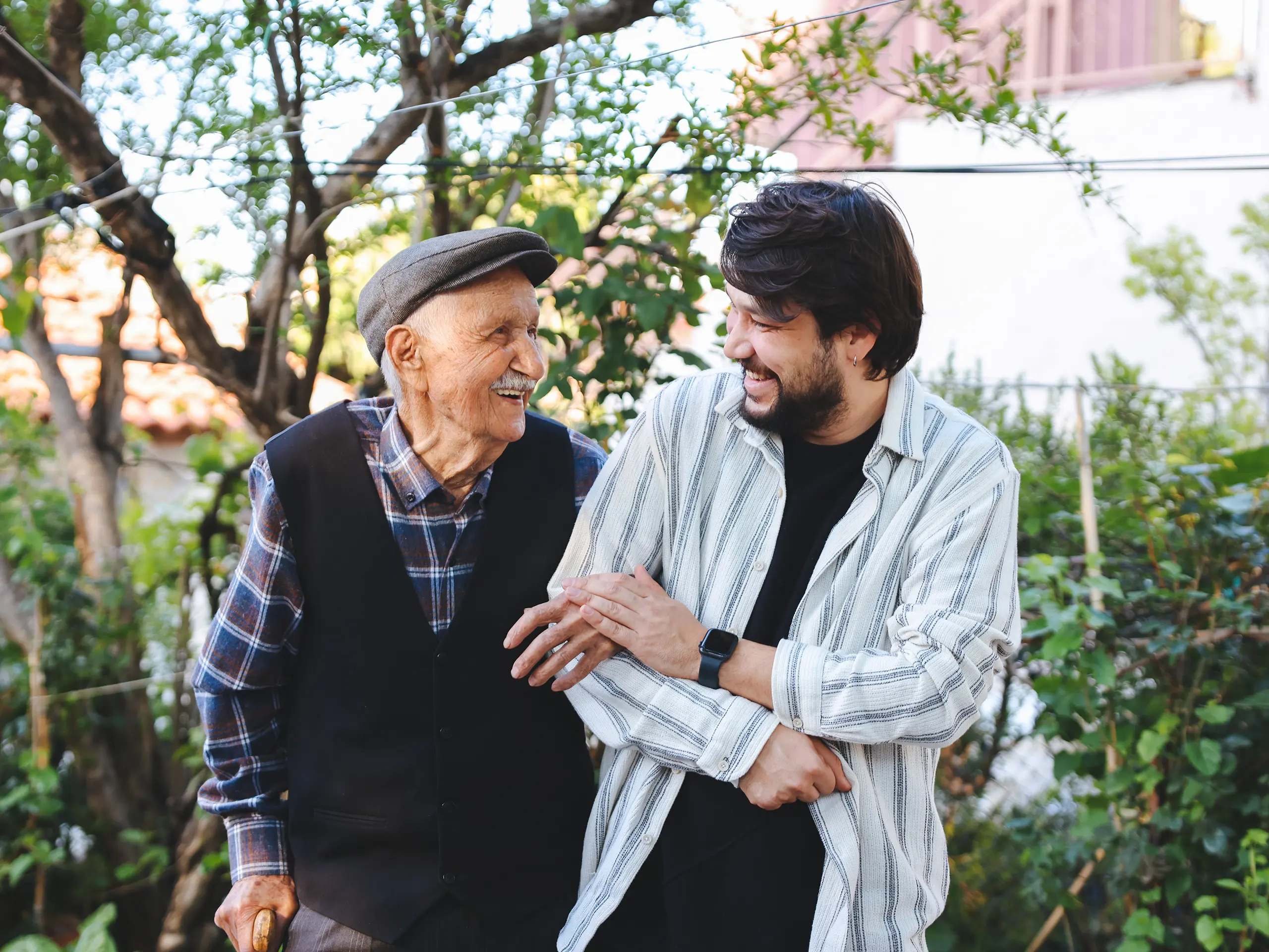 Ein älterer Mann und ein jüngerer Mann gehen gemeinsam durch einen Garten und stützen sich gegenseitig, umgeben von Bäumen und Grün.